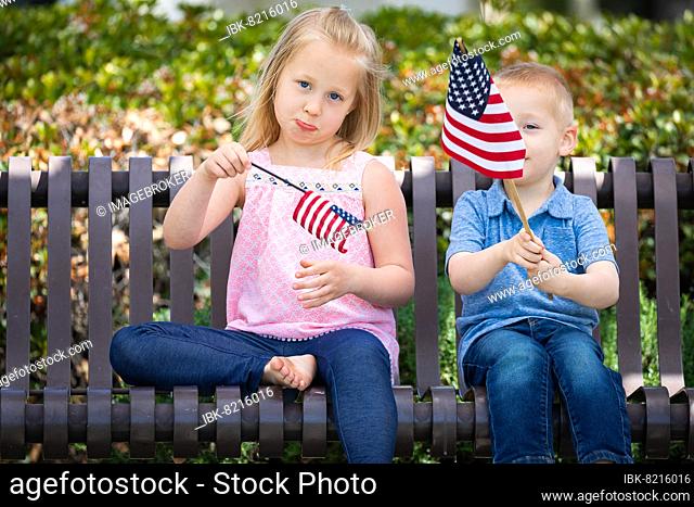 Young sister and brother comparing each others american flag size on the bench at the park