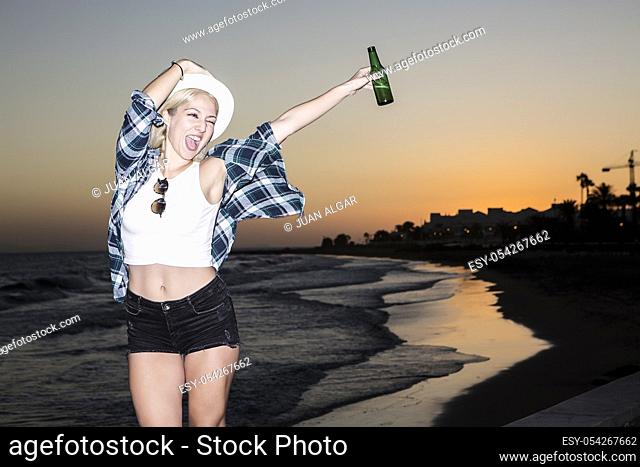 Cheerful young woman hanging out with bottle of beer at seaside in the evening. Lanzarote, Canary Islands, Spain