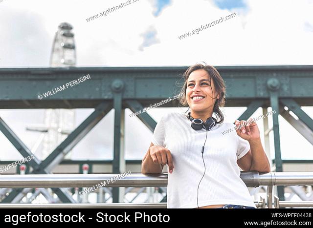 Smiling woman with headphones looking away while leaning on railing at bridge, London, England, UK