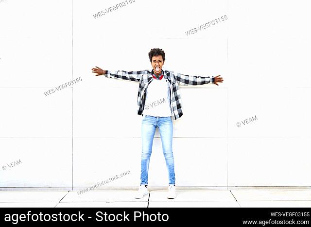 Young female hipster shouting while standing with arms outstretched against white wall