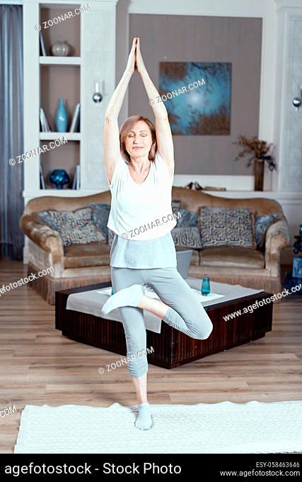 A Blond Woman Aged 50 or 60 Years Engaged in Yoga. She is at Home in the Living Room. The Woman Keeps Fit