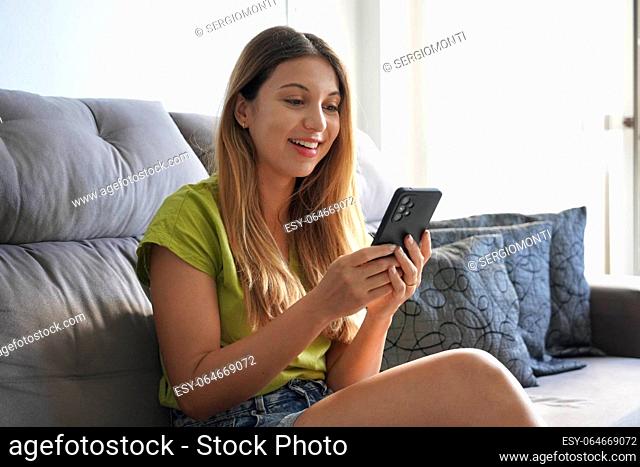 Attractive lively girl resting on cozy sofa with smartphone, share messages to friend in social networks smile enjoy free time on internet