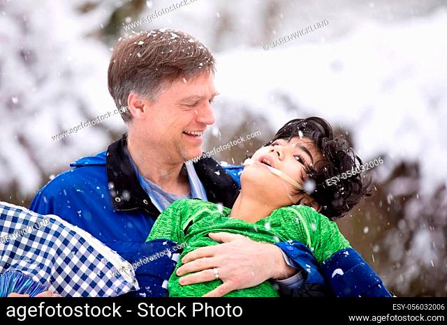 Smiling father holding disabled young son in his arms outdoors during snowfal. Child has cerebral palsy