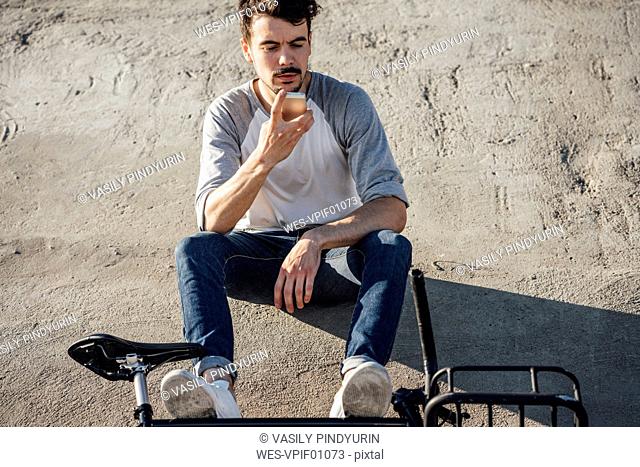 Young man with commuter fixie bike sitting on concrete wall using cell phone
