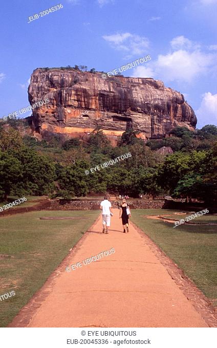View towards huge monolithic rock site of fith century citadel. Also called Lion Rock. Tourist couple on path through gardens in the foreground