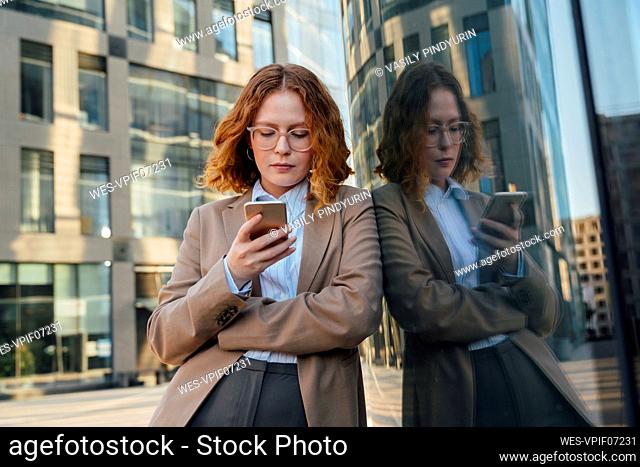 Businesswoman with eyeglasses using smart phone leaning on glass wall