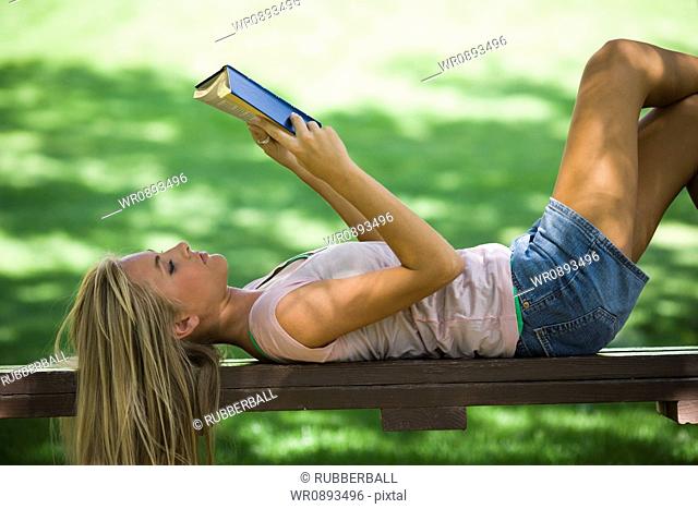 Young woman lying on a park bench and reading a book