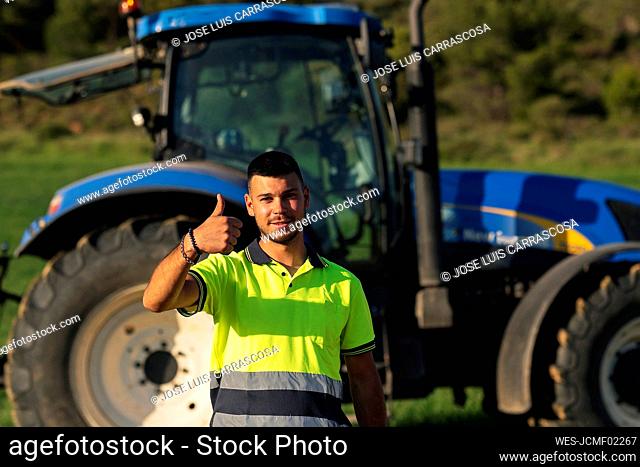 Young farmer gesturing thumbs up in front of tractor