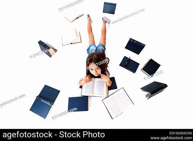 cute little child girl in denim is reading book on the floor with group of books around her on white background. top view