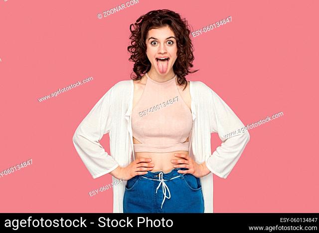 Portrait of funny crazy beautiful brunette young woman with curly hairstyle in casual style standing, hands on waist and tongue out looking at camera