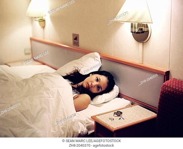 A portrait of a woman lying in a cruise ship hotel bed with her watch and earrings on the bedside table