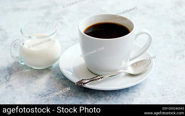 Arrangement of small glass pitcher with fresh milk on marble table with white cup of coffee with silver spoon on saucer