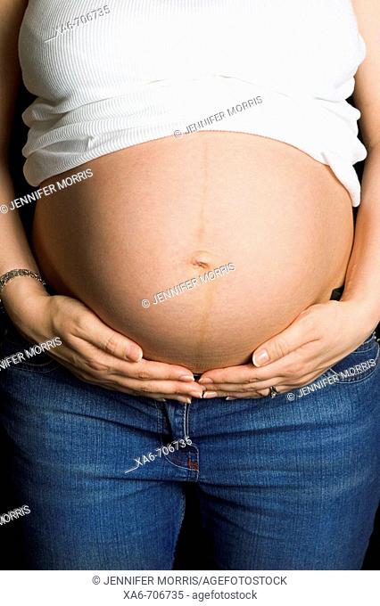 A pregnant woman wearing white t-shirt and blue jeans cradles her bare belly. No face is shown
