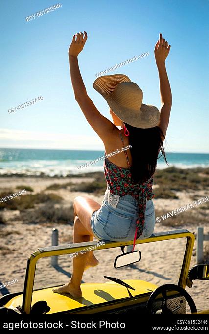 Caucasian woman sitting on beach buggy by the sea wearing straw hat looking toward sea with hands up