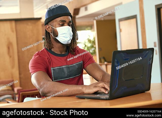 Portrait of young ethnic man wearing red t shirt and knit hat with face mask, sitting at bar in kitchen of downtown loft with laptop computer