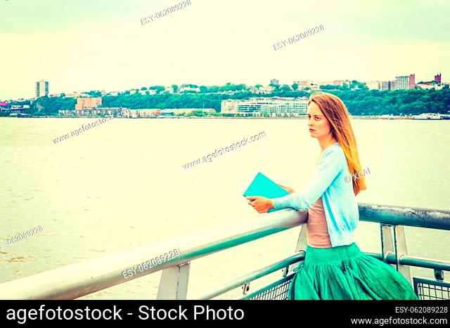 Unforgettable Reading. Wearing light blue cardigan, green skirt, holding book, an American woman standing by metal fence by Hudson River in New York