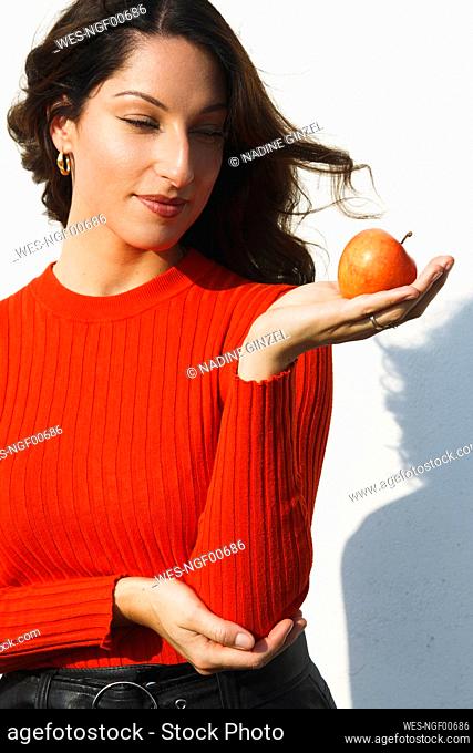 Beautiful woman holding apple while standing against wall on sunny day