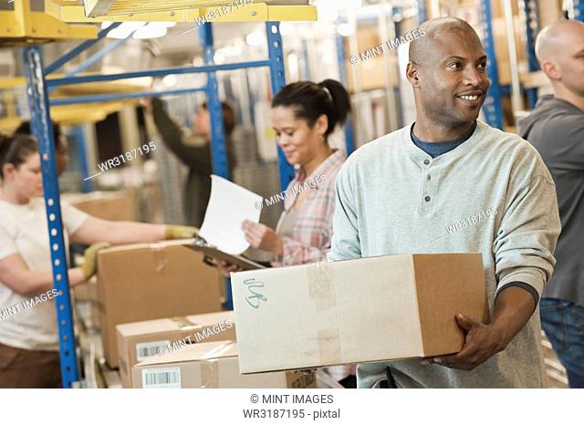 African American warehouse worker and a team of multi-ethnic workers working next to a motorized conveyor of cardboard boxes holding products in a large...