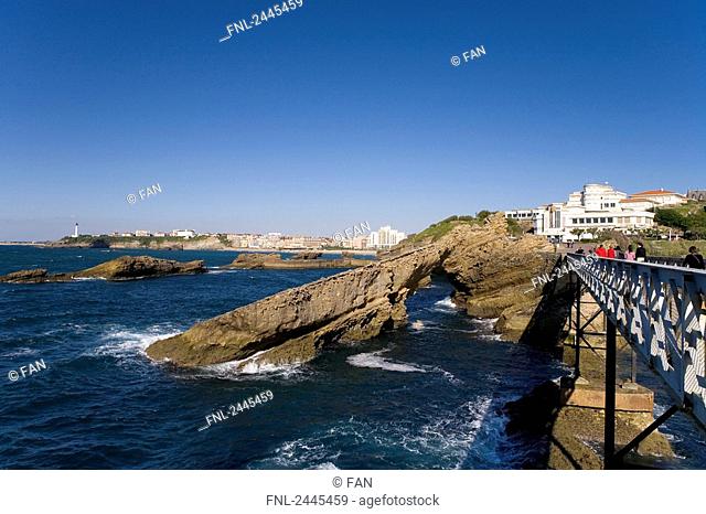 Tourists walking on bridge near natural arch with lighthouse in background, Pointe Saint-Martin, Biarritz, Pyrenees-Atlantiques, Aquitaine, France