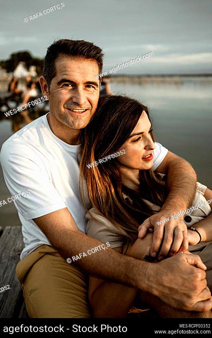 Smiling man embracing woman while sitting at jetty