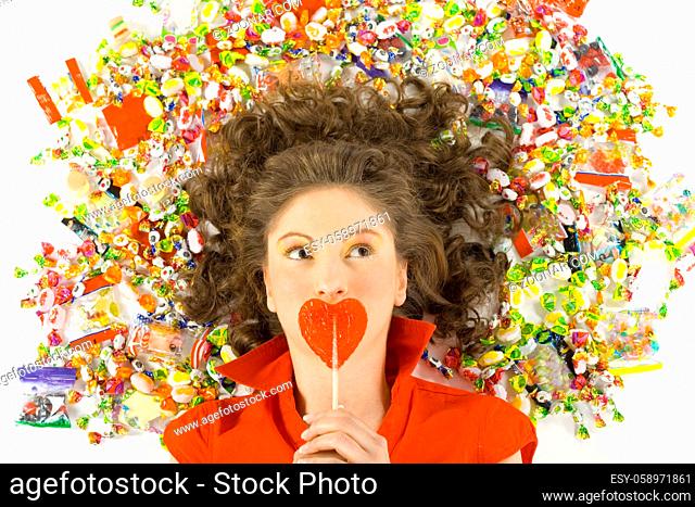 Young, beautiful woman lying on floor among candys with lollypop in hand. Thinking of something, front view. White background