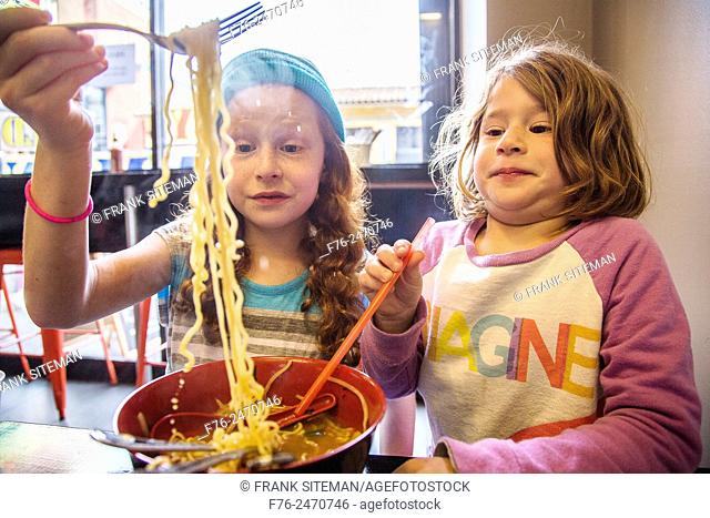 10 year old girl with woolen cap on and her 6 year old sister, eating noodles in a noodle restaurant in Berkeley, CA