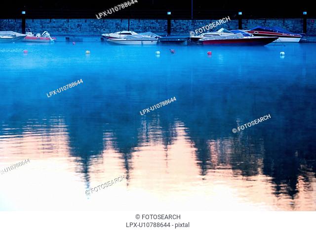 Motorboats moored at Lake Como, , golden light of sunrise and pine trees reflected in water with swirling mists, Lombardy, Northern Italy