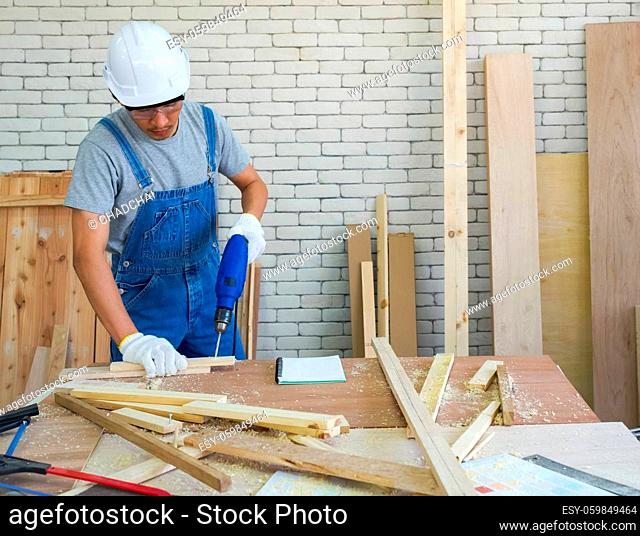 Asian carpentry uses an electric drill to drill the planks as they were prepared at the woodworking facility. Morning work atmosphere in the workshop room