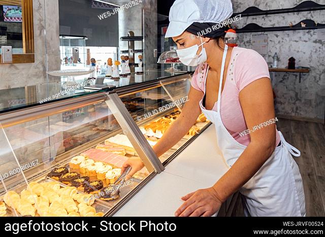 Female confectioner wearing protective face mask arranging pastry in retail display