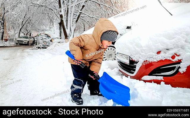 Boy in jacket and grey hat helping to clean up the snow covered red car after snow storm using big blue shovel