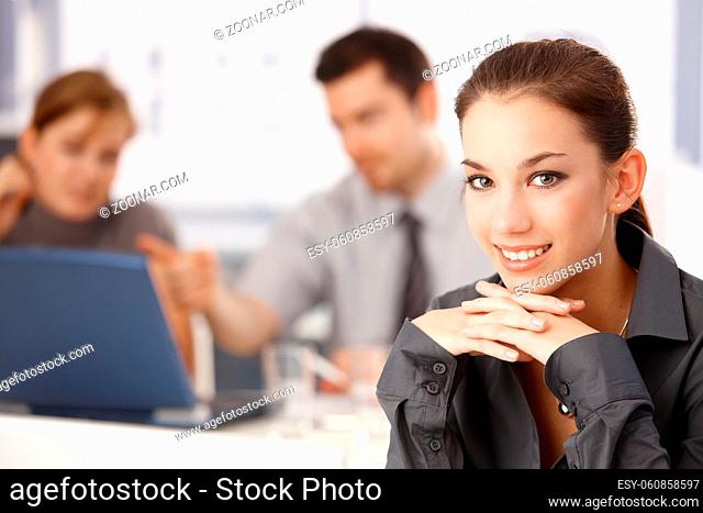 Portrait of attractive businesswoman sitting at meeting table, colleagues working in the background