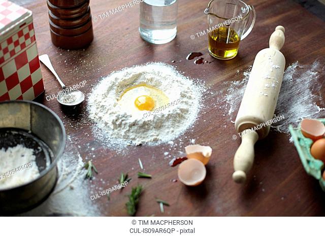 Table with raw egg in center of flour stack and kitchen utensils