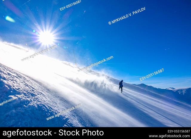 Steep ski slope in windy sunny weather. A lonely unrecognizable snowboarder descends and picks up a lot of snow dust in the backlight of the sun