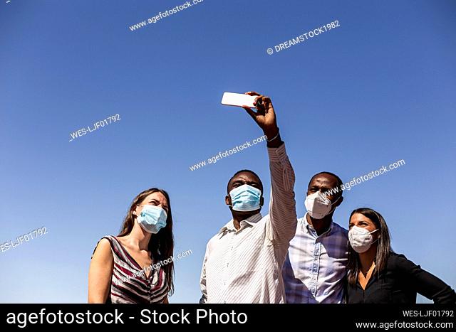 Business professionals taking selfie with protective face masks against clear blue sky on sunny day