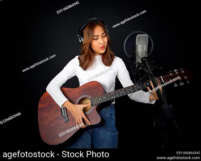 Young asian woman with headphone sing while playing an acoustic guitar in front of black soundproofing wall. Musician producing music in professional recording...