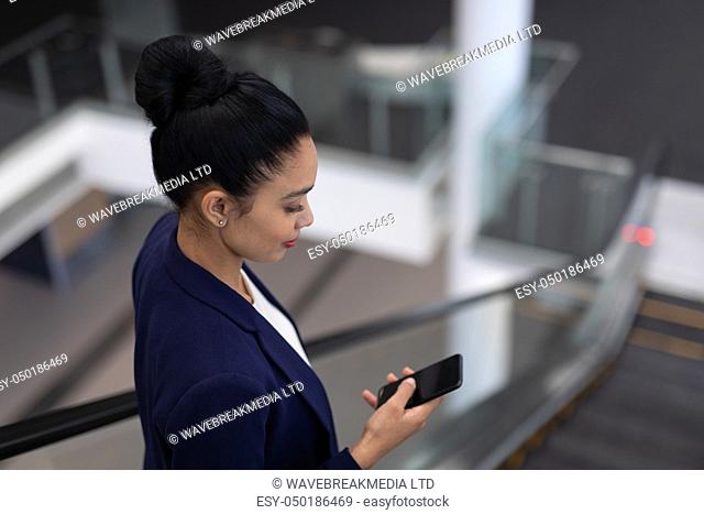 Side view of pretty young mixed-race businesswoman using mobile phone on escalator in modern office
