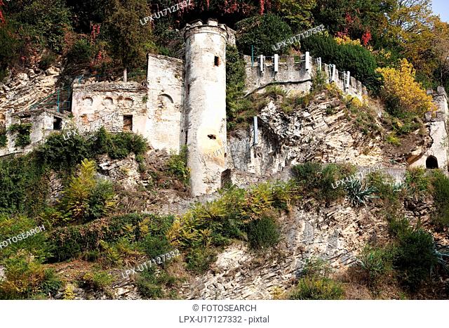 Castle ruins built into hillside with merloned tower and outer walls visible, surrounded by rock, bushes and ferns growing on hillside , Northern Italy