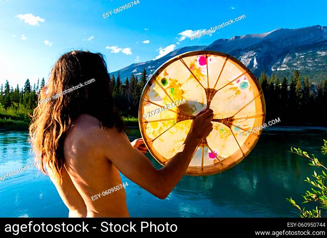 A close up and rear shot of a shamanic drummer playing a sacred indigenous style drum in nature, standing by a calm lake seeking enlightenment