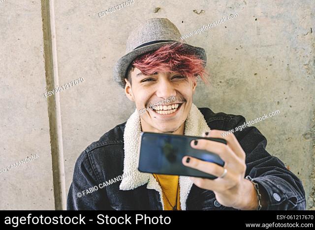 Stylish handsome teenage boy in knit hat and denim jacket with burgundy hair color photographing or taking selfie using mobile phone