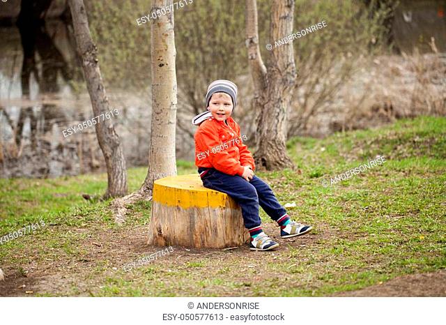 Little boy sitting on a stump in the spring park. Baby boy in orange ja? ket, outdoors