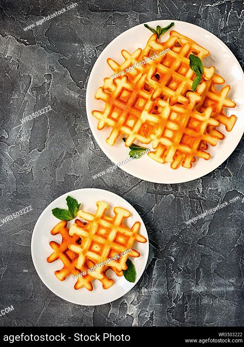 Two plates of Belgian waffles garnished with mint leaves on gray background