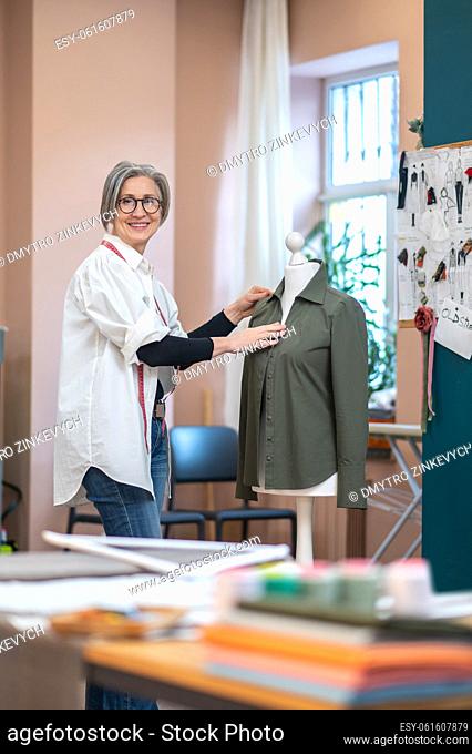 Good result. Happy gray-haired pretty woman in glasses standing near mannequin touching shirt in sewing workshop at daytime