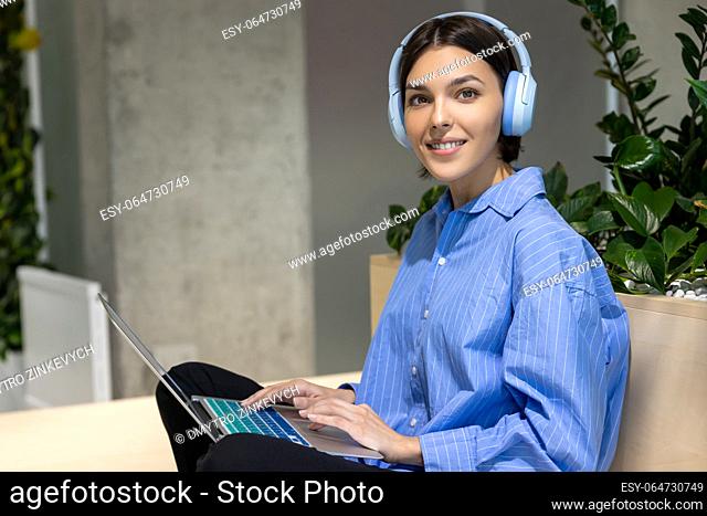 Smiling joyous beautiful young woman in the wireless headphones sitting with the portable computer indoors