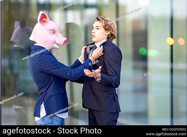 Businessman in pig mask holding collar of male colleague by glass wall