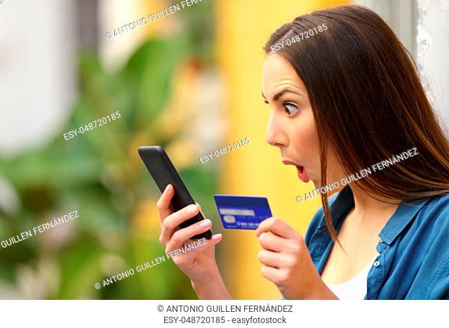 Amazed woman finding surprising offers buying online with smart phone and credit card in a colorful street
