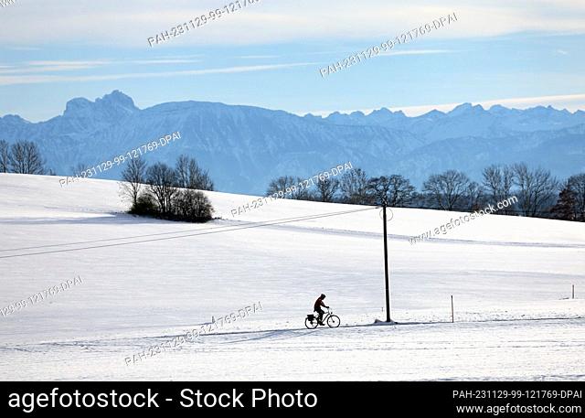 29 November 2023, Bavaria, Ruderatshofen: A cyclist rides through the snowy landscape in the sunshine against the backdrop of the Alps
