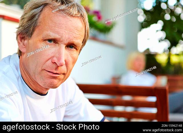 Alter Mann sitzt vor seinem Haus im Sommer und schaut ernst