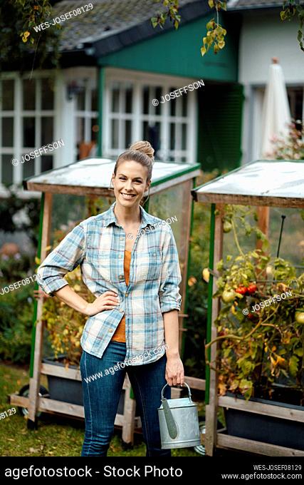 Happy blond woman with hand on hip holding watering can at backyard