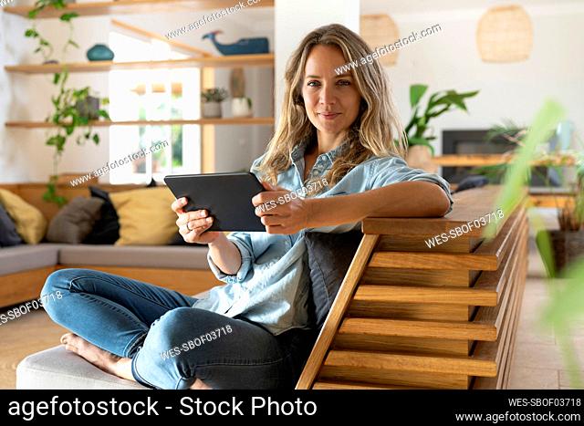 Happy woman holding tablet while sitting with legs crossed on couch in living room