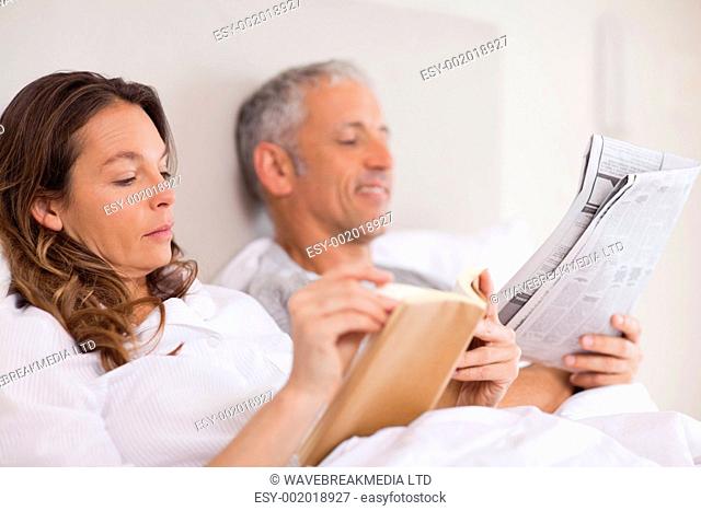 Woman reading a book while her husband is reading a newspaper in their bedroom
